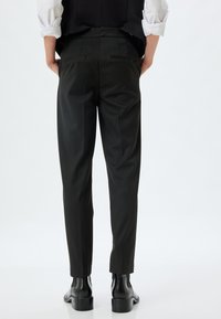 Pantalons noirs ajustés avec une coupe étroite, dotés de deux poches arrière, texture lisse et un éclat subtil, associés à des bottines noires.