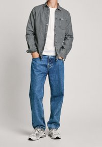Graues Denimhemd über einem weißen T-Shirt, kombiniert mit lockeren blauen Jeans und grauen Sneakers. Das Hemd verfügt über eine Knopfleiste und eine Brusttasche.