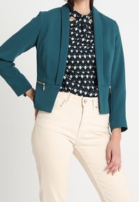 Žena oblečená v modro-zeleném přizpůsobeném sáčku na zip, vzorované černobílé blůze a kalhotách s vysokým pasem v barvě slonové kosti, stojí s rukou na bocích.