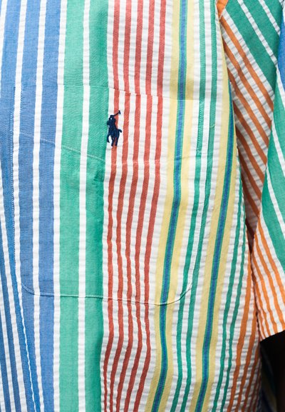 Υφάσμα με πολύχρωμες κάθετες ρίγες και μπλε κεντητό λογότυπο με άλογο και αναβάτη στην τσέπη πουκάμισου.