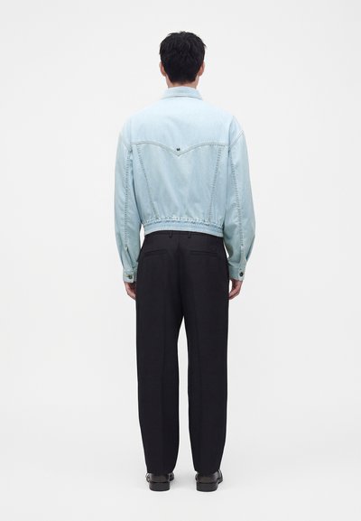 Homme vu de dos portant une veste en jean bleu clair, un pantalon noir et des chaussures noires sur un fond blanc uni.