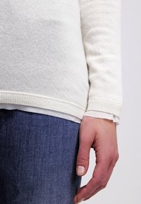 Main droite d'une personne portant un pull en tricot blanc et un jean bleu sur un fond clair uni.