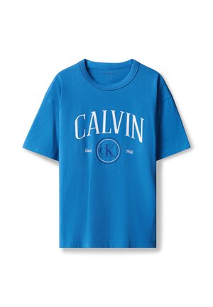 Blondes Kind steht, trägt ein blaues Calvin Klein T-Shirt, hellblaue Jeans und weiße Sneakers vor einem schlichten hellgrauen Hintergrund.