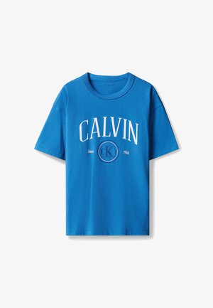 Blond dziecko stoi, ubrany w niebieski t-shirt Calvin Klein, jasnoniebieskie dżinsy i białe tenisówki na jednolitym jasno-szarym tle.