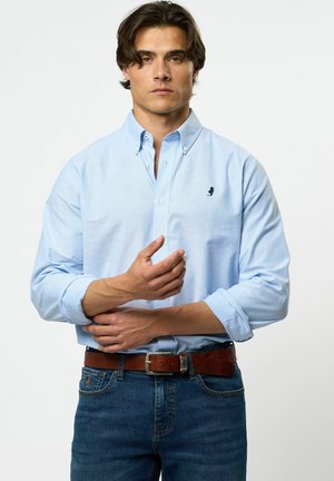 Helleblaue Button-up-Bluse mit Kragen, ausgestattet mit einem kleinen schwarzen Logo auf der Brust, getragen mit dunklen Jeans und einem braunen Gürtel.