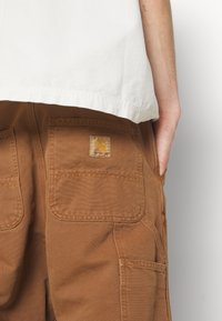 Bruna arbetsbyxor med stora patchfickor, slitstark tyg och en Carhartt-logotyp. Midjan är lätt elastisk för komfort.