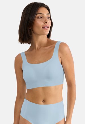 Femme portant un soutien-gorge sans coutures bleu clair et une culotte assortie, regardant sur le côté sur un fond blanc uni.