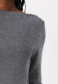 Tessuto grigio, con texture e brillanti metallici. In primo piano, il collo barca di una blusa e la silhouette aderente, che mette in risalto cuciture lisce e maniche lunghe.