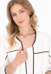Cardigan alb cu detalii negre, design deschis în față și mâneci lungi. Textură moale cu o croială lejeră, purtat peste un top alb.