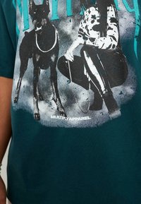 Tyrkysové tričko s grafikou černého psa a osoby v puntíkované košili. Obsahuje opotřebovaný potisk a bílý text "MULTIPLY APPAREL."