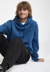 Blauwe katoenen hoodie met een voorzak, verstelbare trekkoord en logoprint. Gecombineerd met zwarte jeans en sneakers. Casual ontwerp.