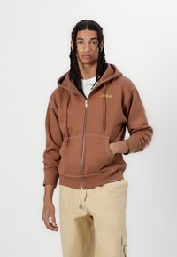 Brown zip-up hoodie with a front pocket, white stitching, and yellow logo; paired with beige pants, the fabric appears soft and casual.