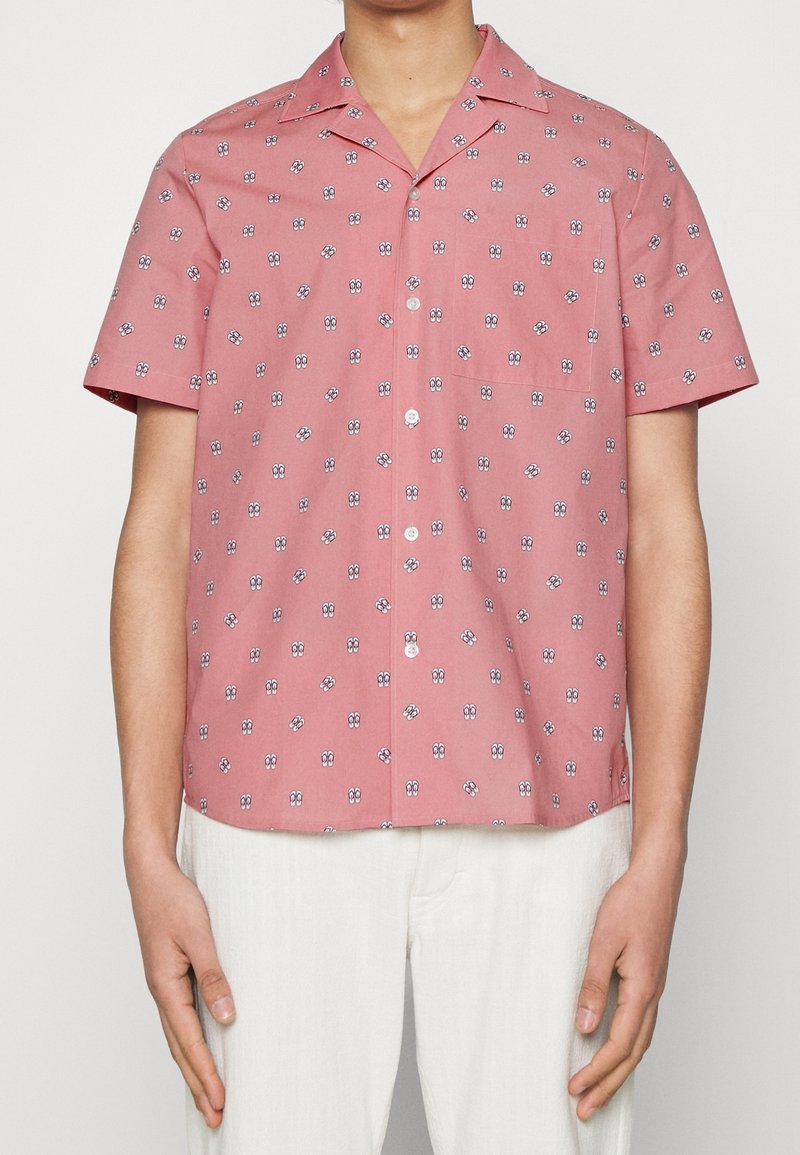 Homme portant une chemise rose à manches courtes avec de petits motifs abstraits en forme d'œil blanc et noir, associée à un pantalon blanc.