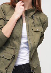 Olivově zelená lehká bunda s vysokým límcem, zipovým předním zapínáním a několika předními kapsami. Má šňůrky v pase a na manžetách.