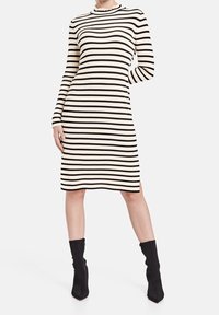 Robe longue à manches longues noire et crème à rayures avec un col haut, texture côtelée, design arrivant aux genoux et fentes sur les côtés. Accompagnée de bottines noires.