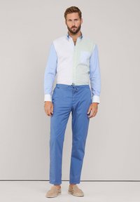 Camisa de botones con secciones verdes y azules, combinada con pantalones azules informales y zapatos de lona beige. Presenta un detalle de bolsillo a rayas.