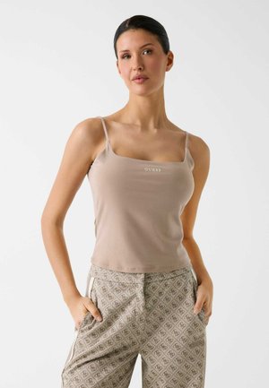 Femme aux cheveux foncés portant un haut beige Guess à fines bretelles et un pantalon beige à motifs, debout les mains dans les poches devant un fond uni.