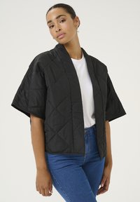 Sort, kviltet jakke med korte ærmer, diamantmønster, åben front, båret over en hvid t-shirt, parret med blå jeans. Glat tekstur.