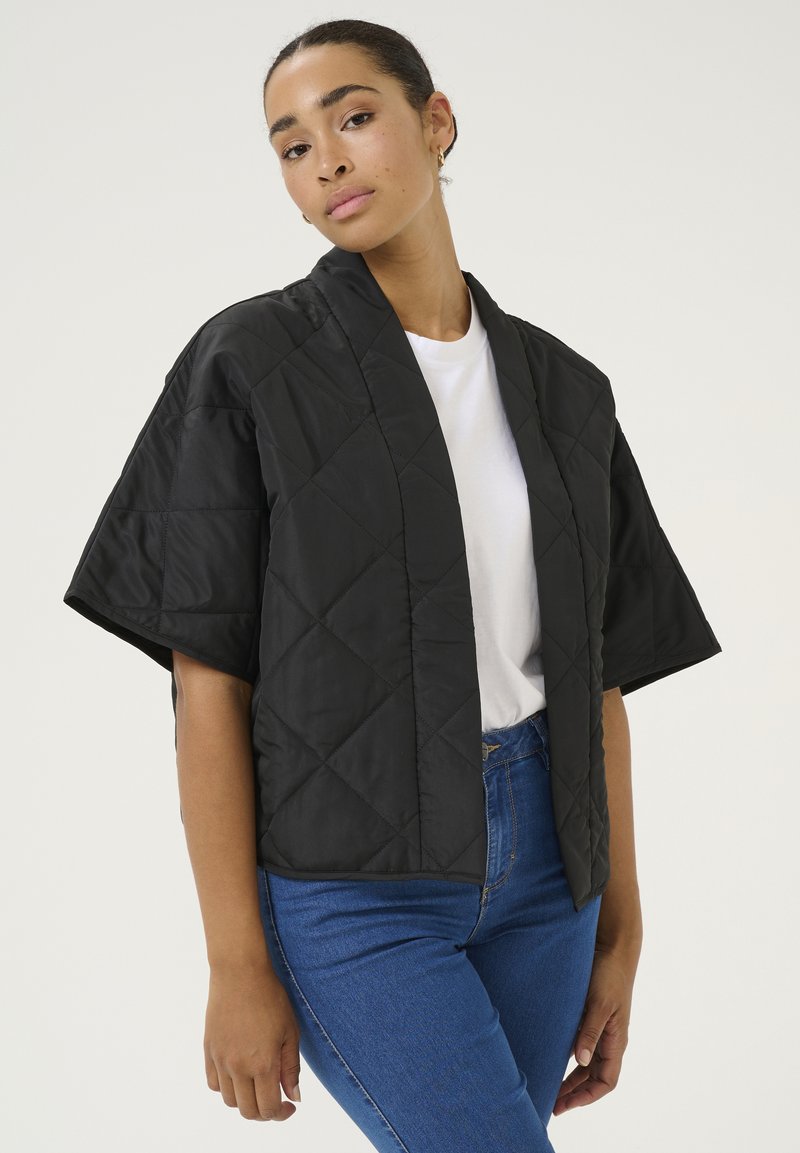 Sort, kviltet jakke med korte ærmer, diamantmønster, åben front, båret over en hvid t-shirt, parret med blå jeans. Glat tekstur.