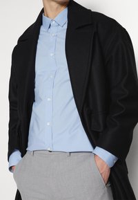 Manteau noir texturé sur une chemise bouton bleu clair, associé à un pantalon gris. Caractéristiques : un large col et des accents de bouton.