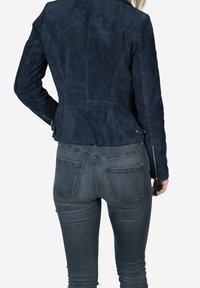 Navy blue suede jacket with fitted design, zippered sleeves, and structured shoulders, paired with gray jeans featuring back pockets.