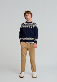 Granatowy sweter z geometrycznym kremowym wzorem, beżowe spodnie i białe trampki. Sweter ma okrągły dekolt i ściągacze.