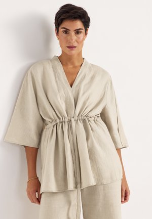 Kvinde med kort mørkt hår iført beige hørkjole med brede ærmer og snørre i taljen, stående foran en hvid baggrund.