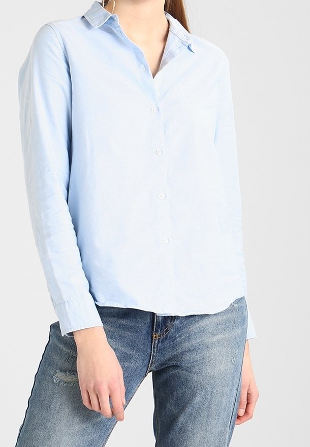 Personne portant une chemise bleu clair à manches longues boutonnée et un jean bleu, debout les mains détendues le long du corps.
