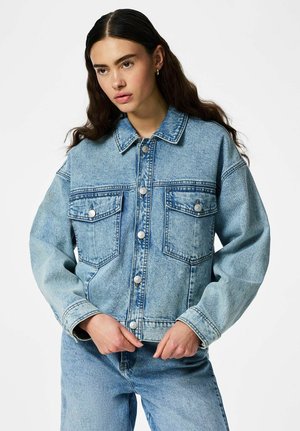 Vrouw met lang donker haar die een lichtblauwe spijkerjasje en bijpassende spijkerbroek draagt, staand met de handen bij de taille.