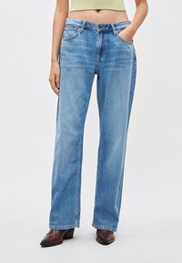 Svijetloplave traperice s visokim strukom, širokim nogavicama, pet džepova i suptilnim izblijedjenjima, uparene s tamnim čizmama špicastog vrha.