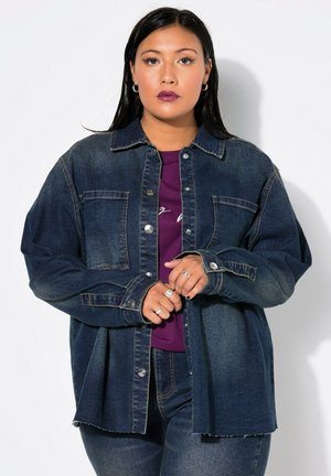 Veste en denim bleu foncé avec un devant à boutons, deux poches poitrine et une coupe décontractée. Superposée sur un haut violet, avec un ourlet effiloché.