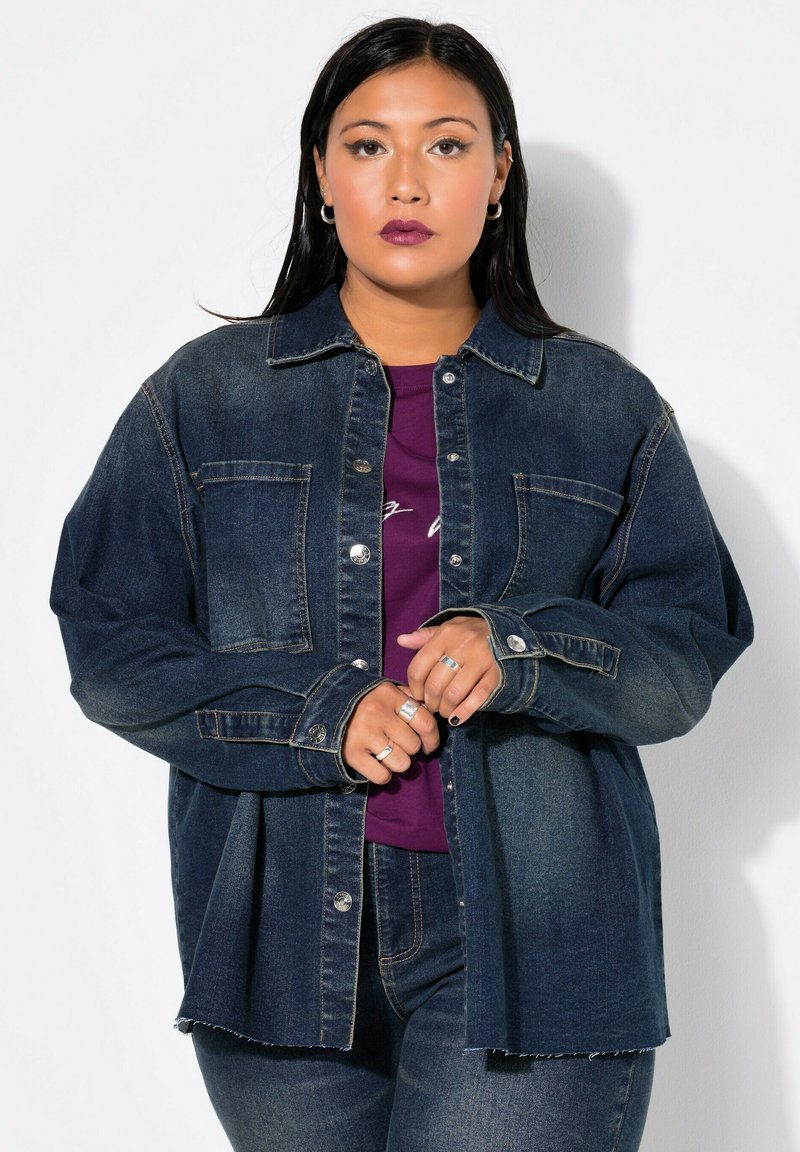 Chaqueta de mezclilla de color azul oscuro con un cierre de botones en el frente, dos bolsillos en el pecho y un corte relajado. Superpuesta sobre una blusa morada, que presenta un dobladillo deshilachado.