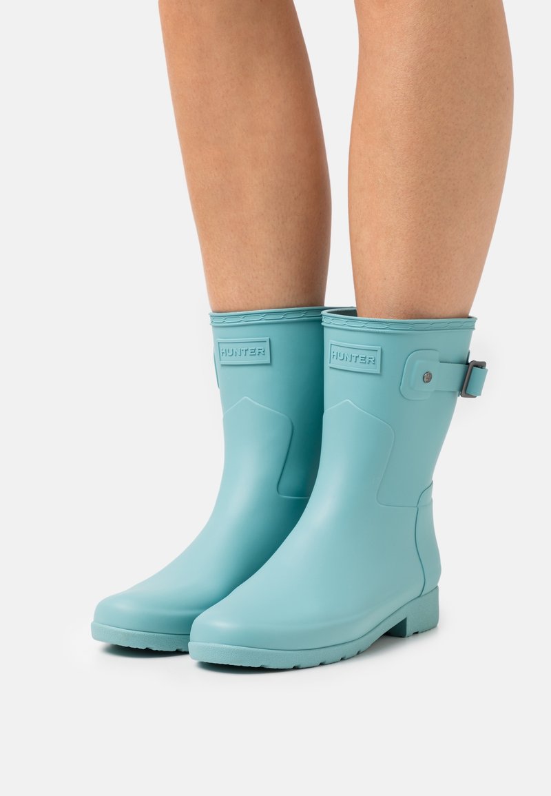 Bottes de pluie en caoutchouc bleu clair avec un bout arrondi, une semelle texturée, un petit talon et un détail de boucle réglable sur le côté. Marque Hunter visible.