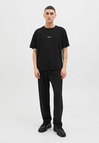 Sort bomulds t-shirt med løs pasform, rund hals og subtilt frontlogo. Båret med sorte bukser og sorte sko. Simpelt design.