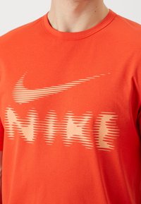 Homme portant un t-shirt Nike orange vif avec un logo déformé et un design swoosh sur la poitrine.
