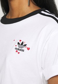 Bílý bavlněný tričko s černými lemy a ohrnutými rukávy. Na přední straně se nachází vyšívané černé logo Adidas obklopené červenými a růžovými srdíčky.