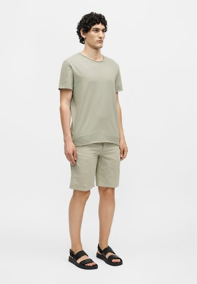 Νέος άνδρας που φοράει μπεζ κοντομάνικη μπλούζα, αντίστοιχα σορτς και μαύρα ανοιχτά πέδιλα, στέκεται μπροστά σε λευκό φόντο.