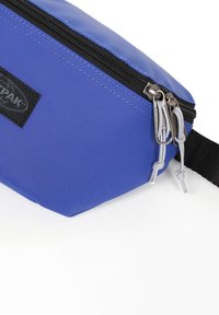 Custodia in tessuto blu con superficie testurizzata. Presenta una cerniera nera e linguette grigie, etichetta del marchio sul lato. Design compatto.