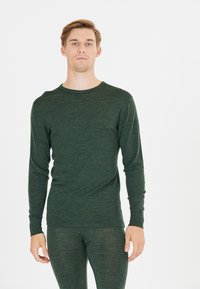Jonge man die naar voren kijkt in een donkergroene lange mouw top en bijpassende slim fit broek tegen een witte achtergrond.