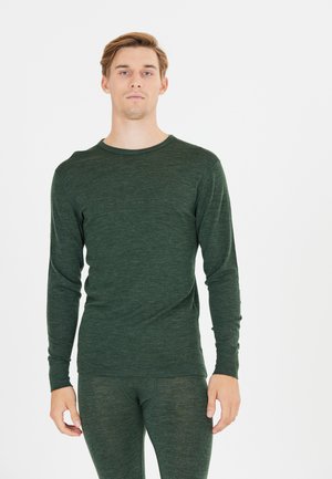 Jonge man die naar voren kijkt in een donkergroene lange mouw top en bijpassende slim fit broek tegen een witte achtergrond.