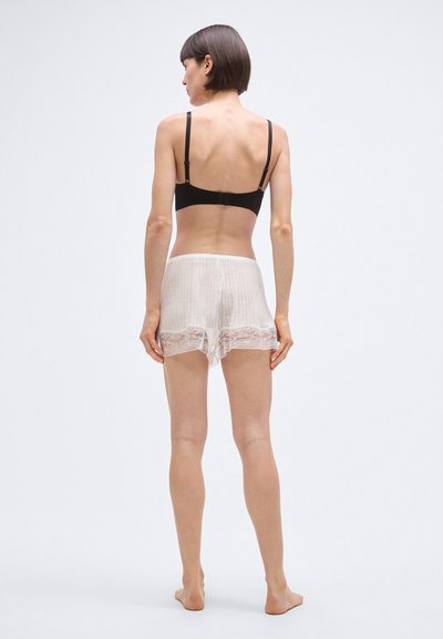 Črni bralet z nastavljivimi naramnicami, oblečen s prosojnimi belimi čipkastimi kratkimi hlačami s črtastim vzorcem in valovitim robom.