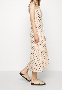 Vestido de lunares en beige con puntos marrones, con tirantes finos y una silueta fluida, acompañado de sandalias de tira en color tostado.