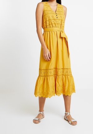 Vestido midi amarillo sin mangas con bordado de ojales, botones en el corpiño, cintura anudada y bajo festoneado, llevado con sandalias plateadas con tiras en el tobillo.