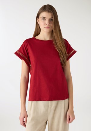 Giovane donna con lunghi capelli castani che indossa una maglietta rossa a maniche corte con dettagli di frange e pantaloni beige, in piedi contro uno sfondo semplice.