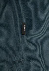 Blue corduroy fabric with vertical ribbing, featuring a black label with "CHASIN'" sewn near the seam. Soft texture and muted color.