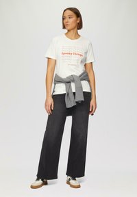 Witte t-shirt met rode en oranje tekst "Spooky Things", gecombineerd met donkergrijze wijde jeans en een grijze gebreide trui om de taille geknoopt.