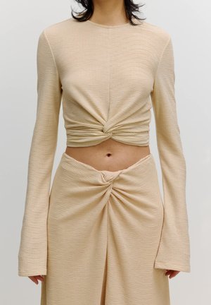 Beige, geribbelde stoffen set bestaande uit een lang getailleerd shirt met een gedraaide knoop in de taille en een bijpassende rok met een gestrikt detail aan de voorkant.