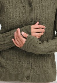 Oliivivihreä neulecardigan, jossa on teksturoitu kuvio, ribbireunat ja viisi nappia. Lähikuva, joka korostaa käsiä hihoissa.