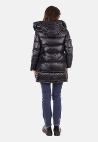 Giacca lunga nera imbottita con cappuccio, caratterizzata da una texture lucida, motivo a righe orizzontali e polsini elasticizzati; indossata con jeans blu e stivali neri.