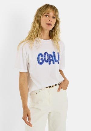 Mujer con cabello rubio y ondulado, que lleva pantalones blancos y una camiseta blanca con la palabra "GOAL!" en azul con lentejuelas, de pie con una mano en el bolsillo.